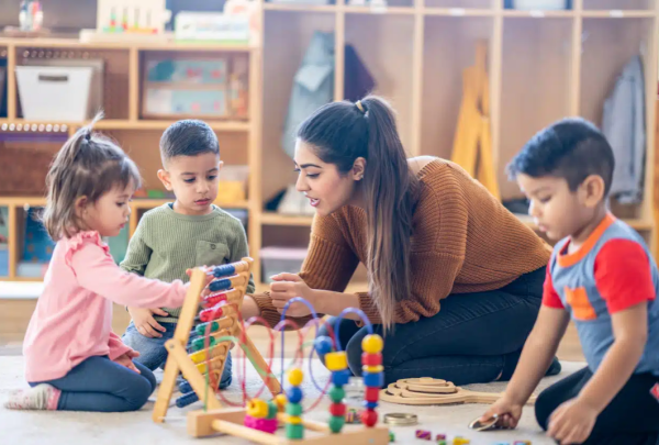Een kinderbegeleider zit met drie kindjes op de grond in een kinderopvang en begeleidt hen bij hun spel.